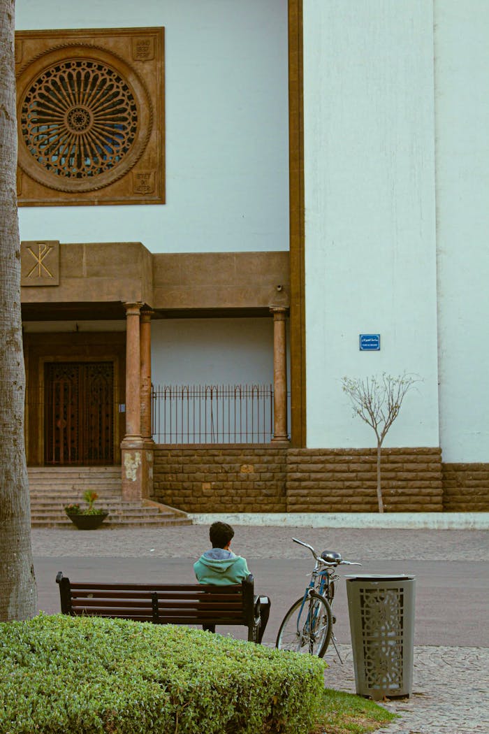 services-img A young man sits on a bench in Rabat, Morocco, near a historic building with a bicycle.