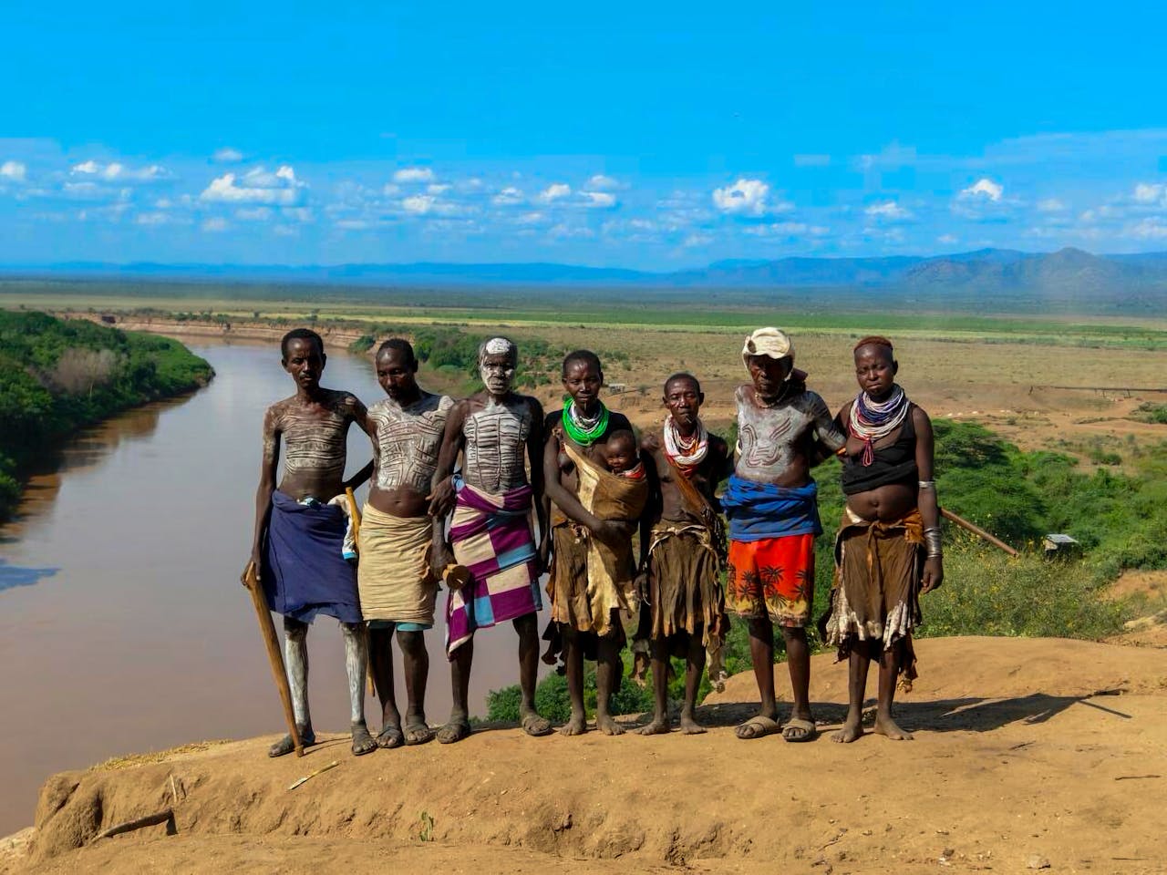 gallery-2 Indigenous group in traditional attire poses by a scenic river landscape under clear blue skies.